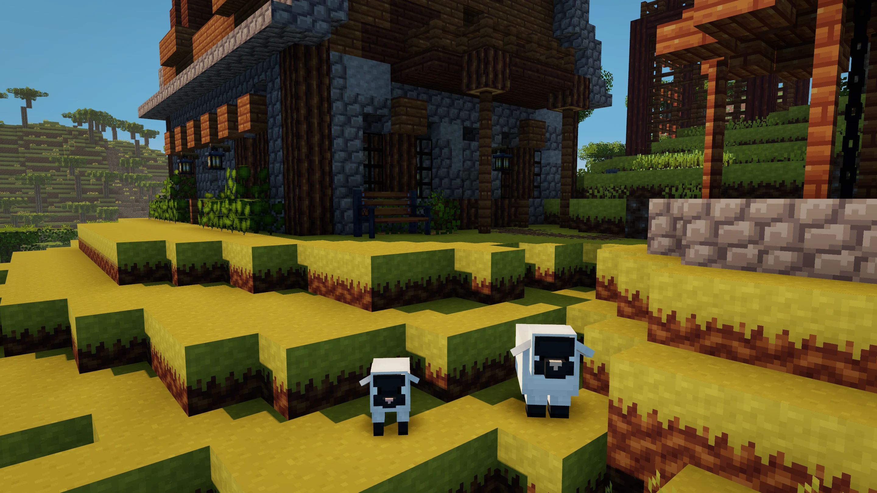 A sheep and lamb on a grassy terrace beside a covered well and a stone-and-timber house